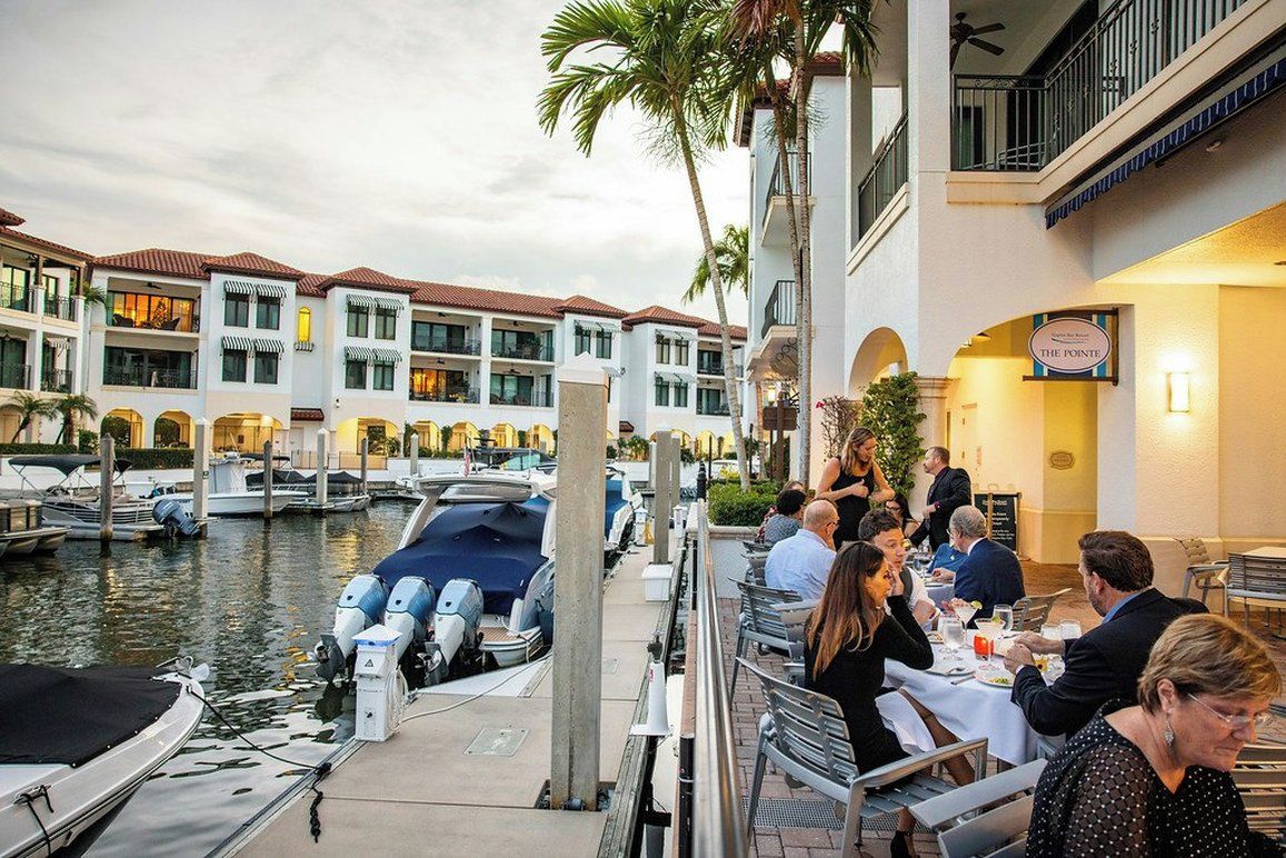 Un puerto deportivo con barcos amarrados, un restaurante con comensales y un edificio de hotel con balcones.