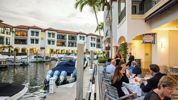 Un puerto deportivo con barcos amarrados, un restaurante con comensales y un edificio de hotel con balcones.