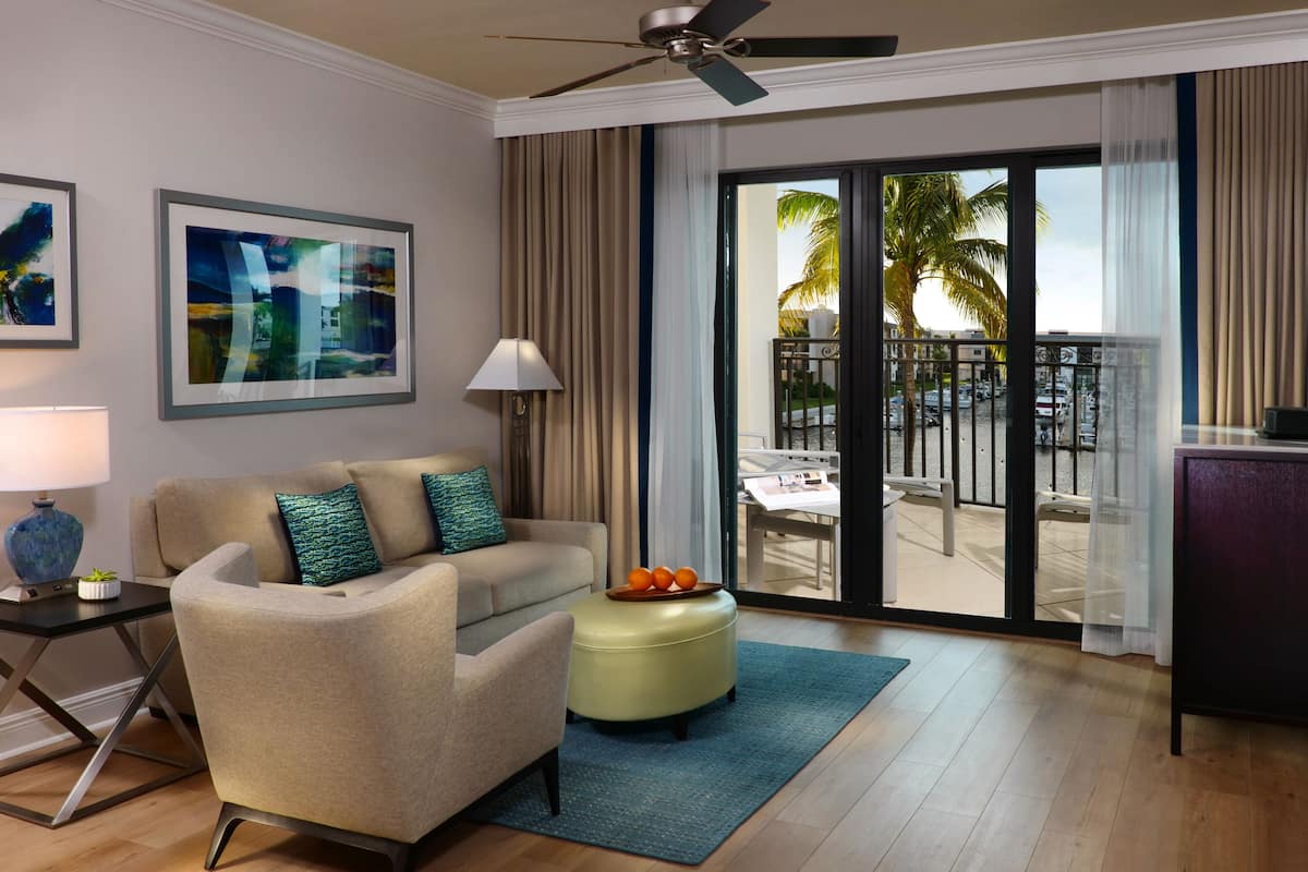 A hotel room with a sofa, ottoman, and a view of a balcony with palm trees.
