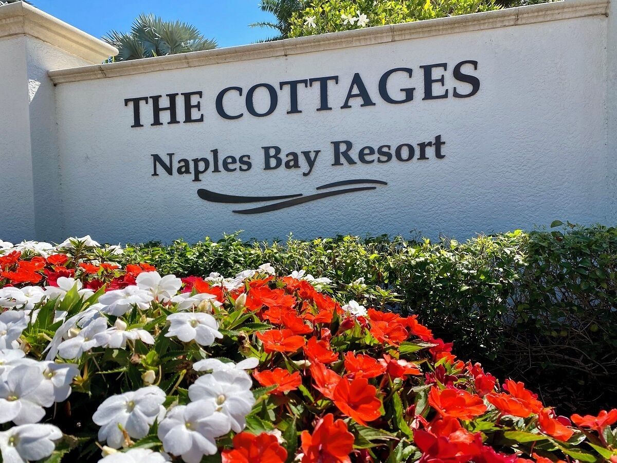Skilt med teksten "The Cottages" ved Naples Bay Resort, omgivet af en have med røde og hvide blomster.
