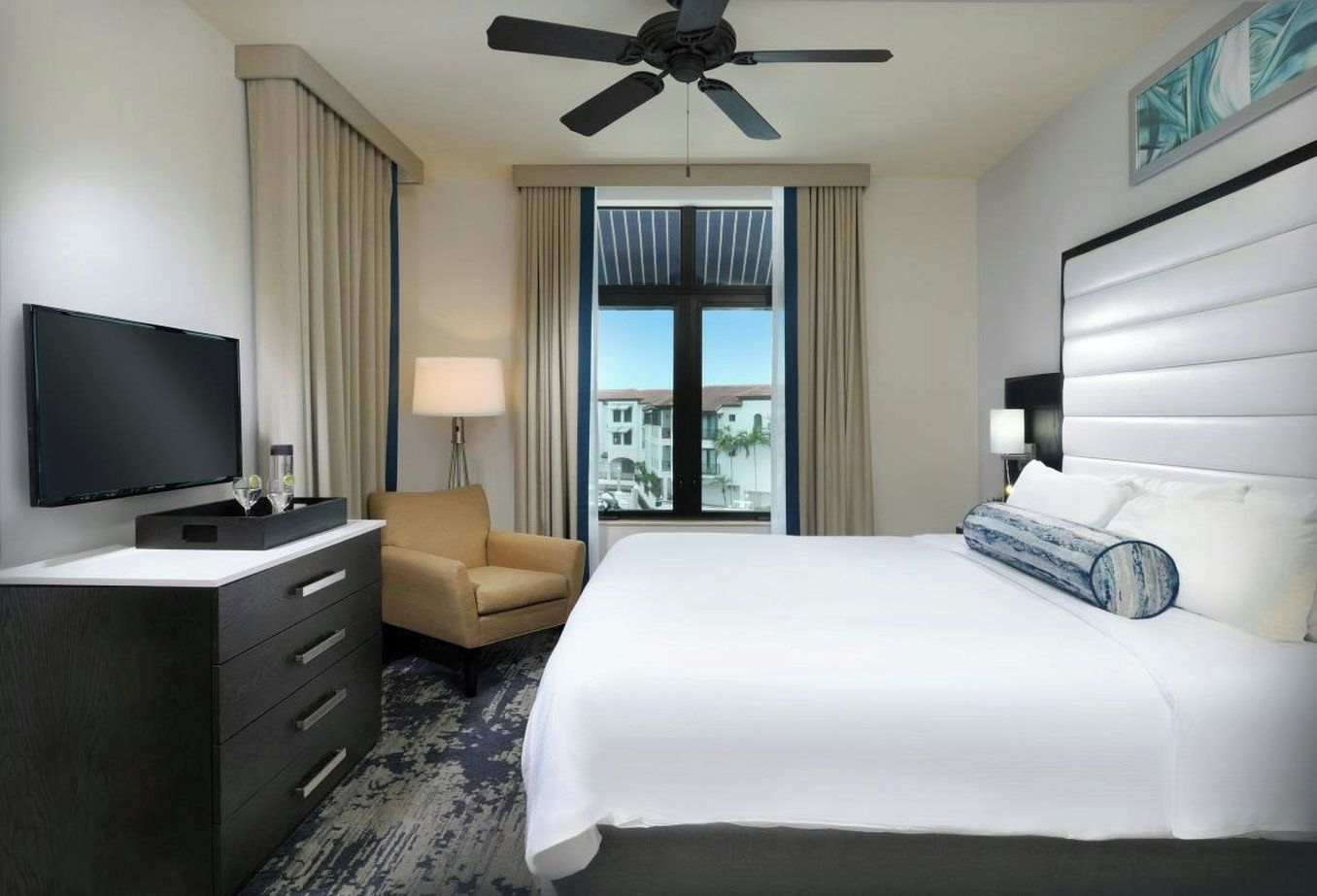 Ein Hotelzimmer mit Bett, Fernseher, Schreibtisch, Stuhl und Deckenventilator.