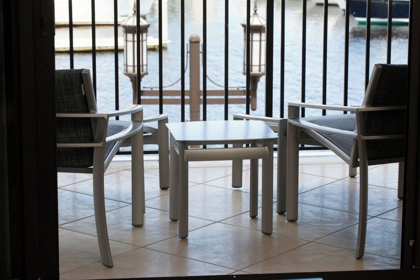 Un balcón con sillas y una mesa de metal, con vista a una masa de agua con barcos y una costa lejana.