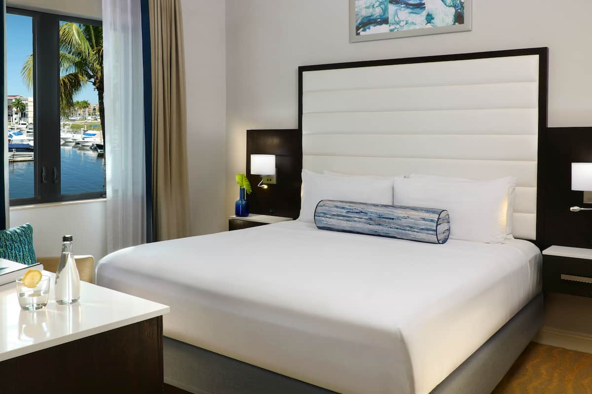 A hotel room with a large bed, two bedside tables with lamps, a small table with a glass and bottle, a window with a view of boats and palm trees, and a painting on the wall.