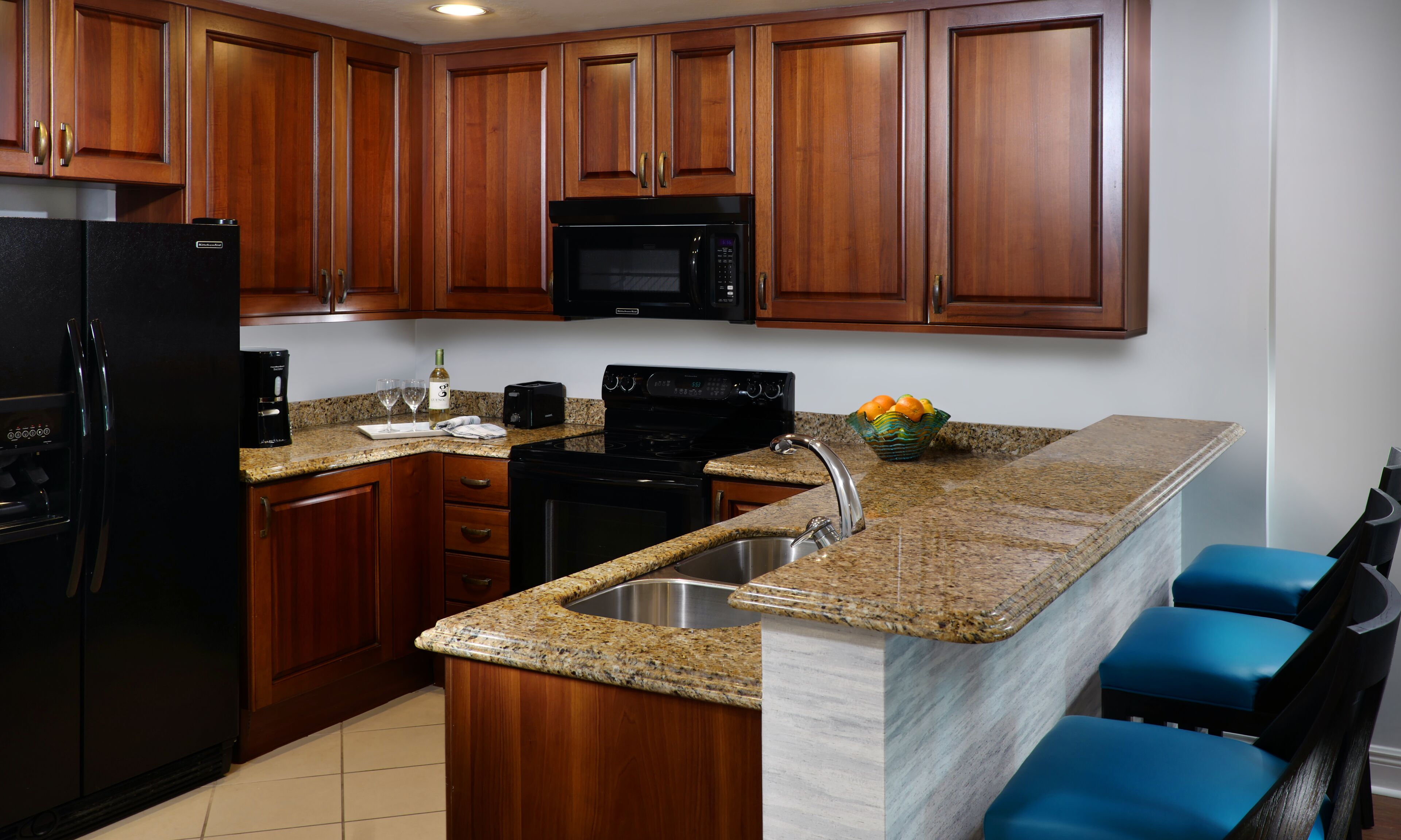A kitchen with wooden cabinets, a black refrigerator, a microwave, a stove, and a granite countertop. There are two blue chairs and a bowl of fruit on the counter.