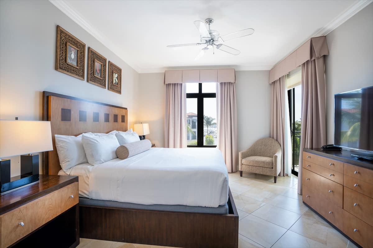A hotel room with a large bed, bedside lamps, a ceiling fan, a TV, a chair, and a window with curtains.