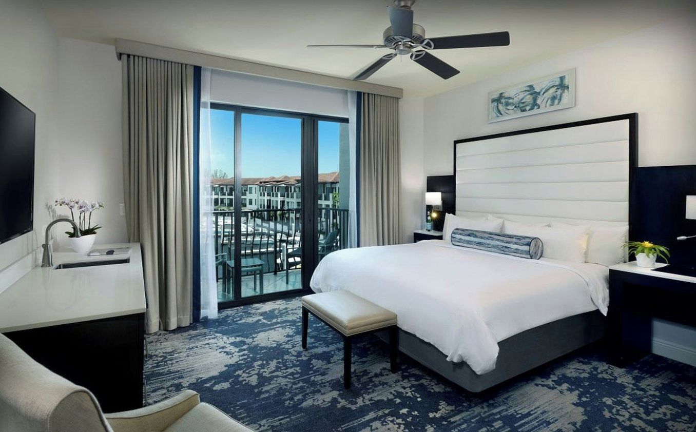 A hotel room with a large bed, a desk, a chair, a ceiling fan, a window with a view, and a painting on the wall.