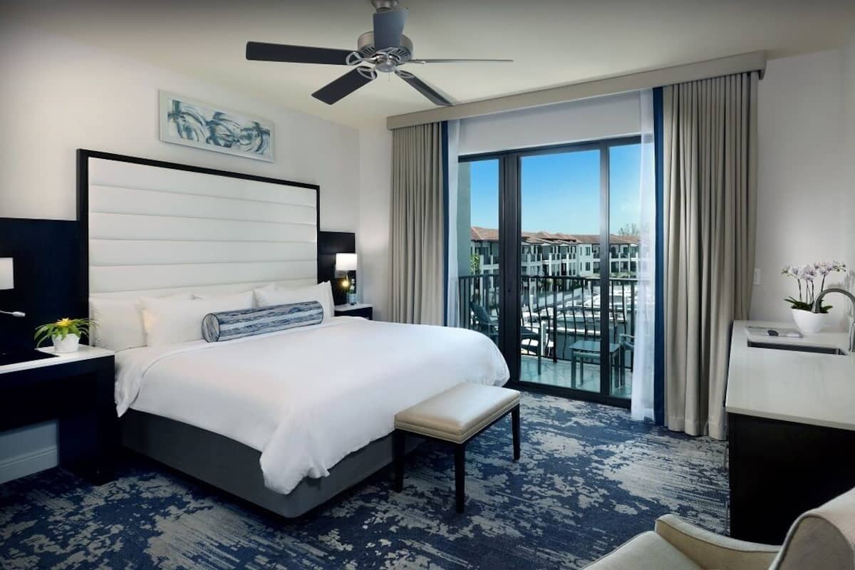A modern hotel room with a large bed, a ceiling fan, a balcony with a view of buildings, and a desk with a plant.