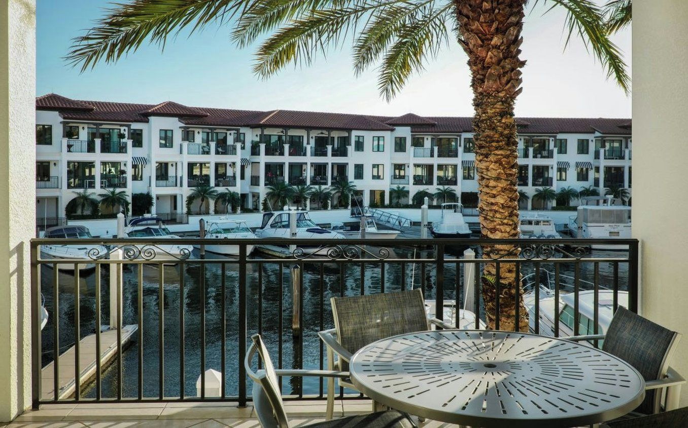 A balcony with a metal railing, a round table, and chairs, overlooking a marina with boats and a multi-story building with balconies.