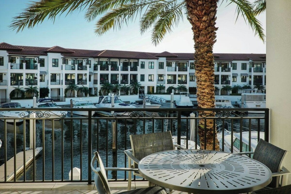 A balcony with a metal railing, a round table, and chairs, overlooking a marina with boats and a multi-story building with balconies.