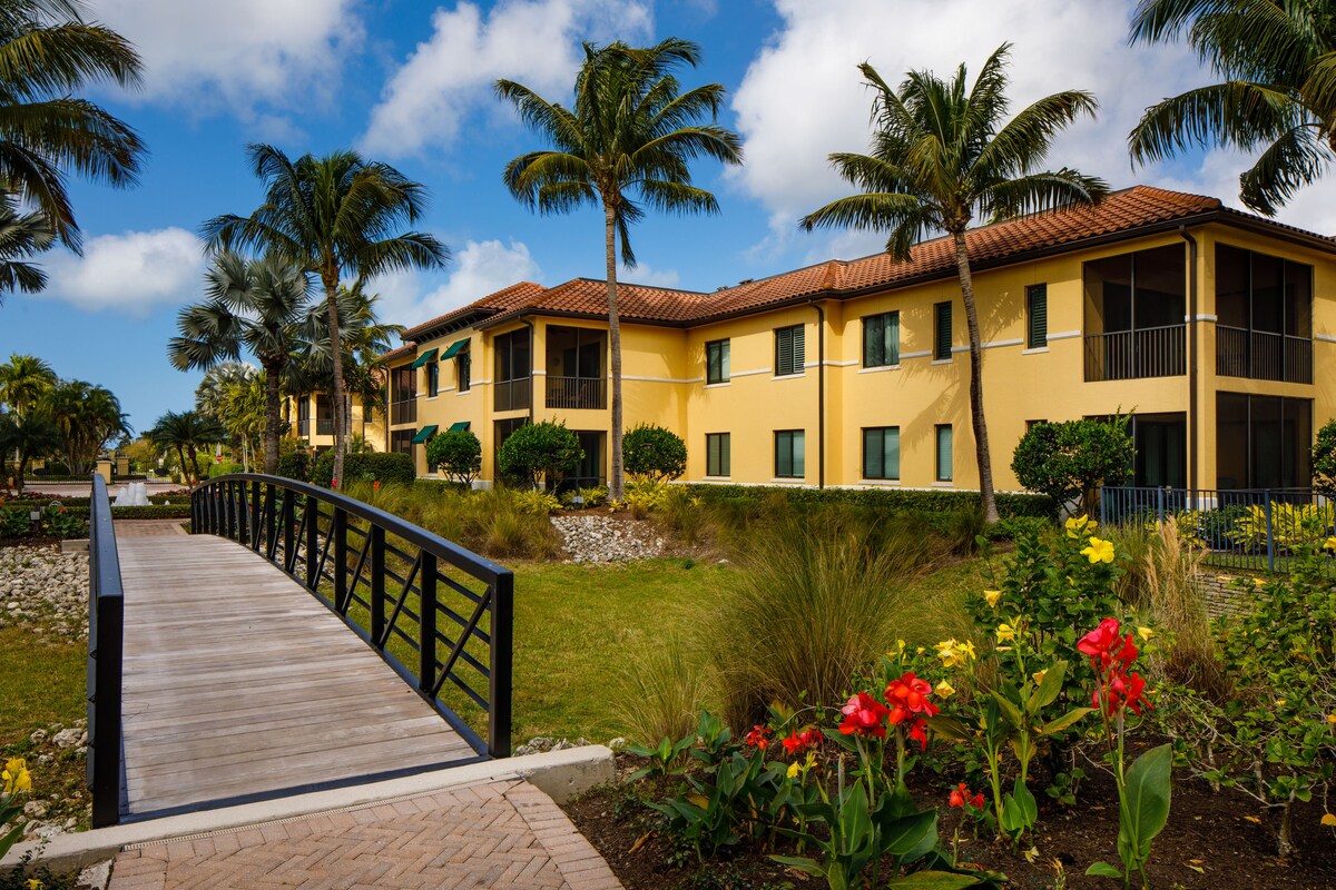 A yellow building with a red roof, surrounded by palm trees and a wooden bridge.