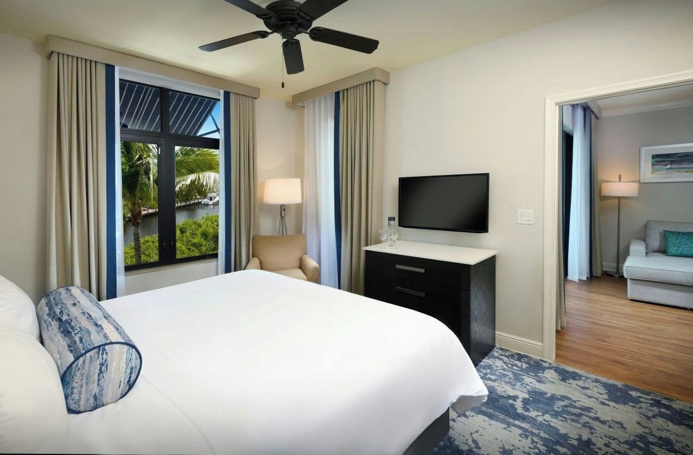 Quarto de hotel com cama, TV de tela plana, escrivaninha com luminária e vista para piscina e vegetação.