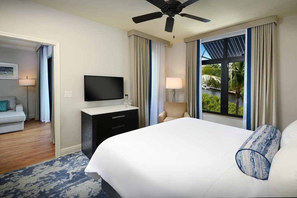 A hotel room with a bed, a TV, a desk with a lamp, a chair, and a window with a view of greenery.