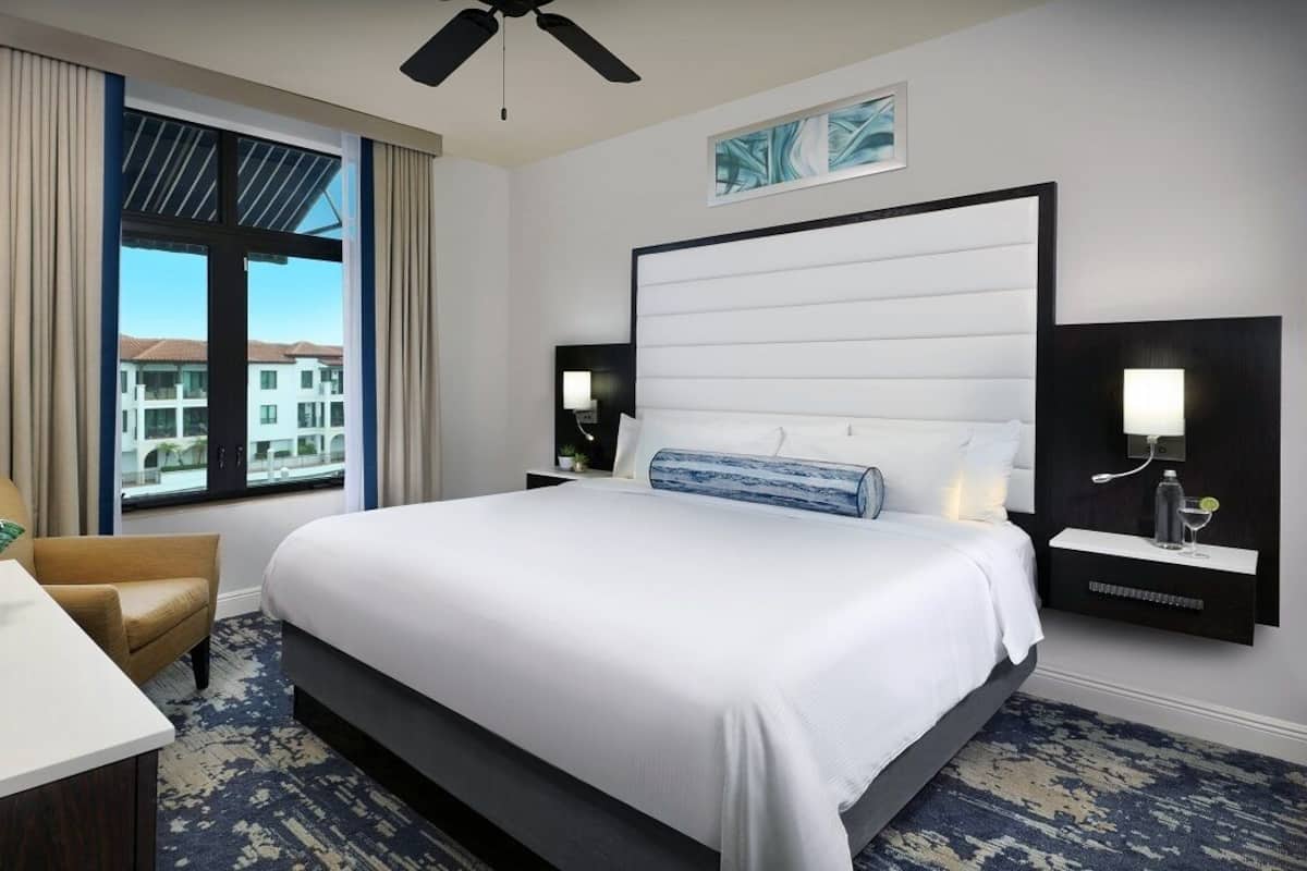 A hotel room with a large bed, a desk, a chair, a window with a view of buildings, and a ceiling fan.