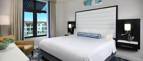 A hotel room with a large bed, a desk, a chair, a window with a view of buildings, and a ceiling fan.