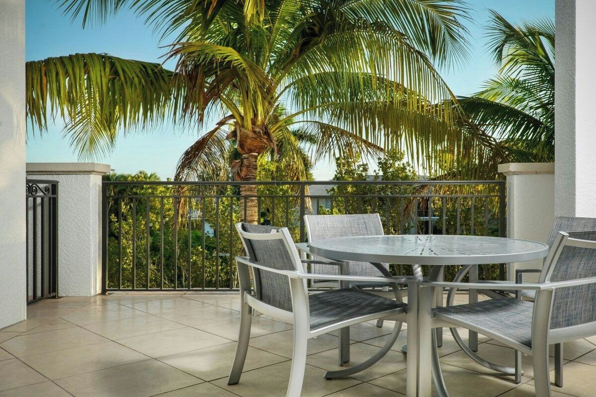 A balcony with a round table and chairs, surrounded by a metal railing, palm trees, and a clear sky.