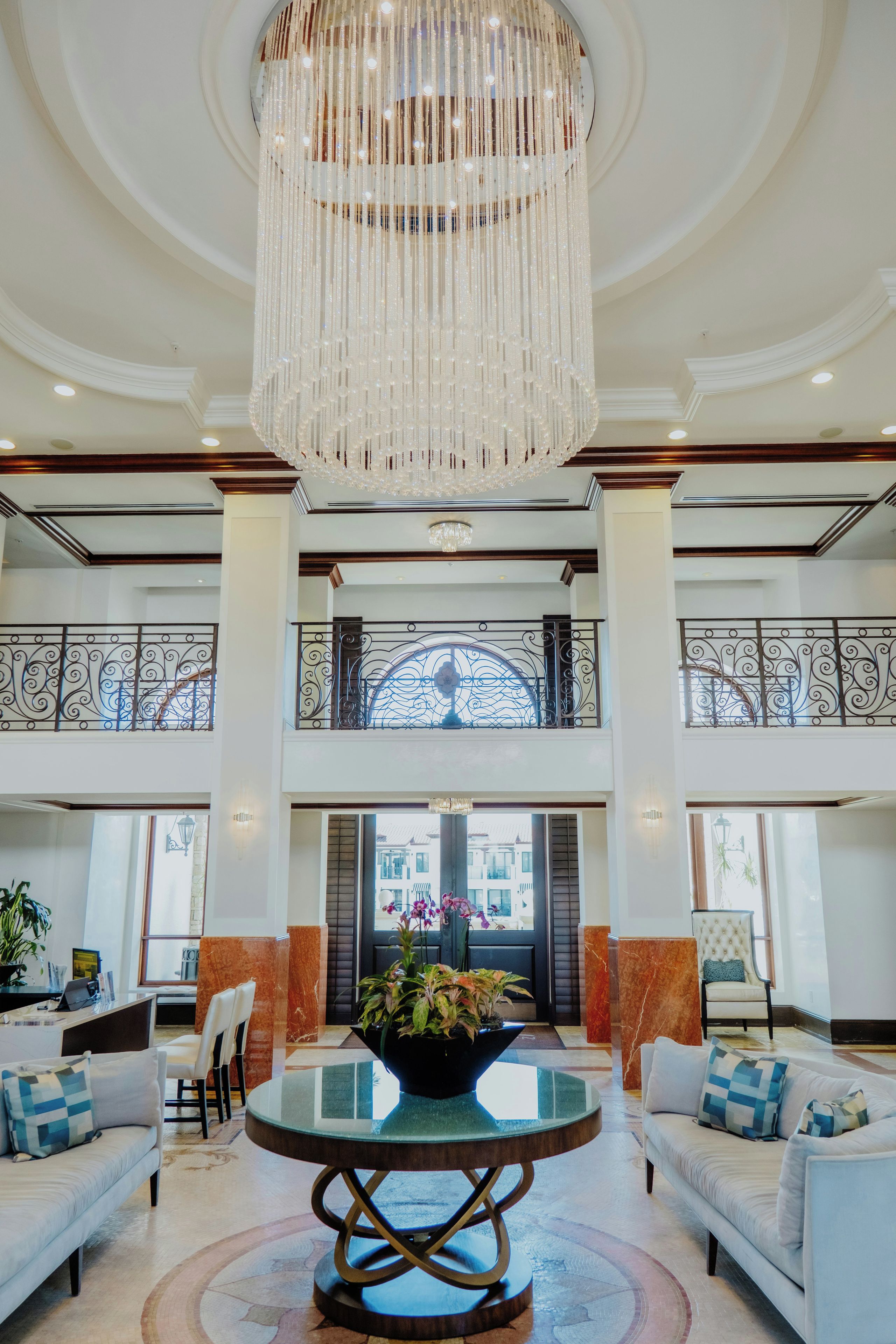 A spacious lobby with a grand chandelier, a round glass table with a plant centerpiece, and a view of the building's exterior through large windows.