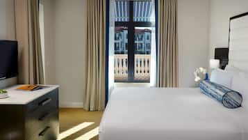 A hotel room with a bed, a desk with a TV, a window with curtains, and a view of a building outside.