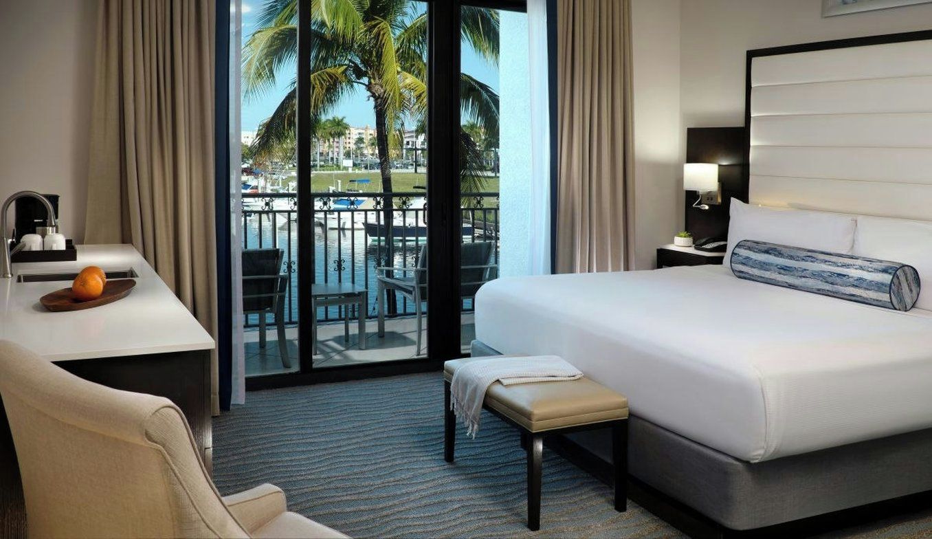 Habitación de hotel con cama, escritorio con lámpara, silla, banco y vista a una piscina y palmeras.