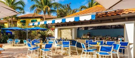 An outdoor bar area with blue and white striped awnings, white metal chairs, and a stone wall backdrop.