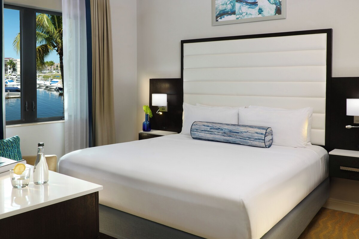 A hotel room with a large bed, two bedside tables with lamps, a small table with a glass and bottle, a window with a view of boats and palm trees, and a painting on the wall.