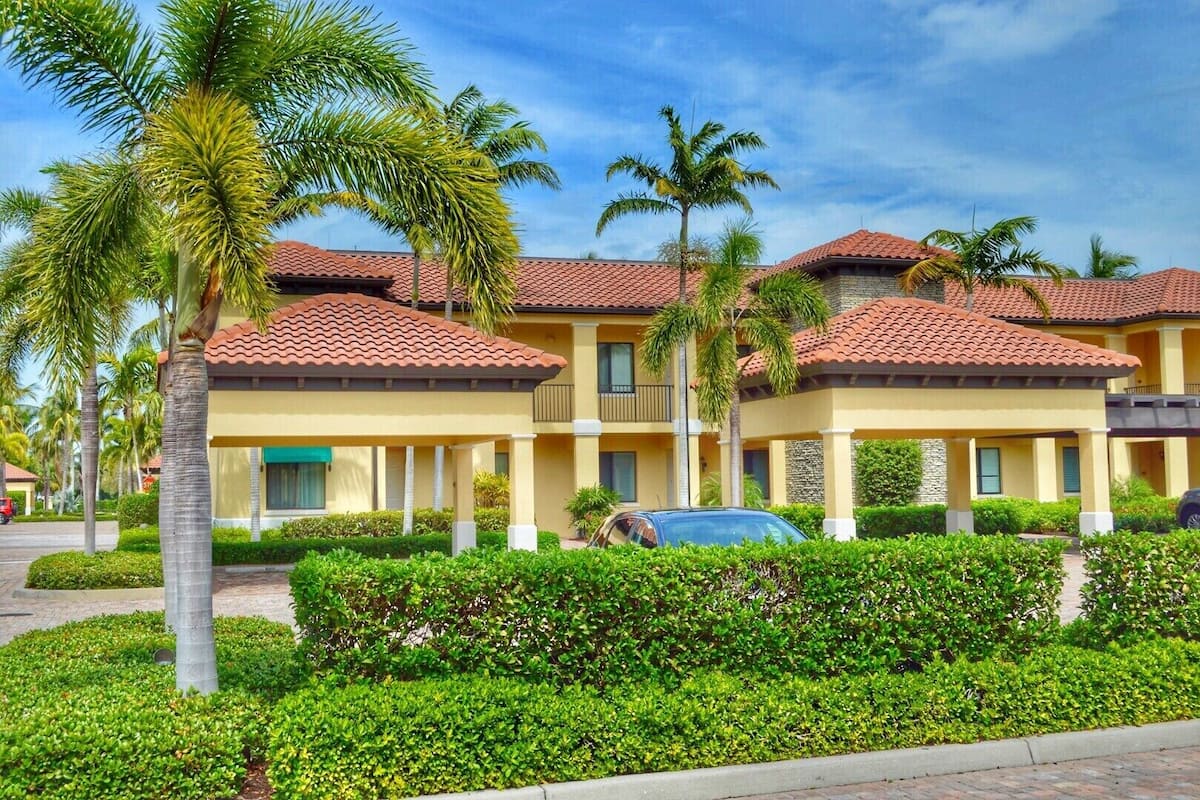 A multi-story hotel with a red-tiled roof, yellow walls, and a central courtyard with palm trees and well-maintained hedges.