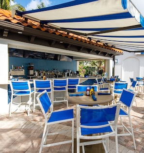 An outdoor bar area with white and blue chairs, a round table, and a bar counter with various bottles and a coffee machine.