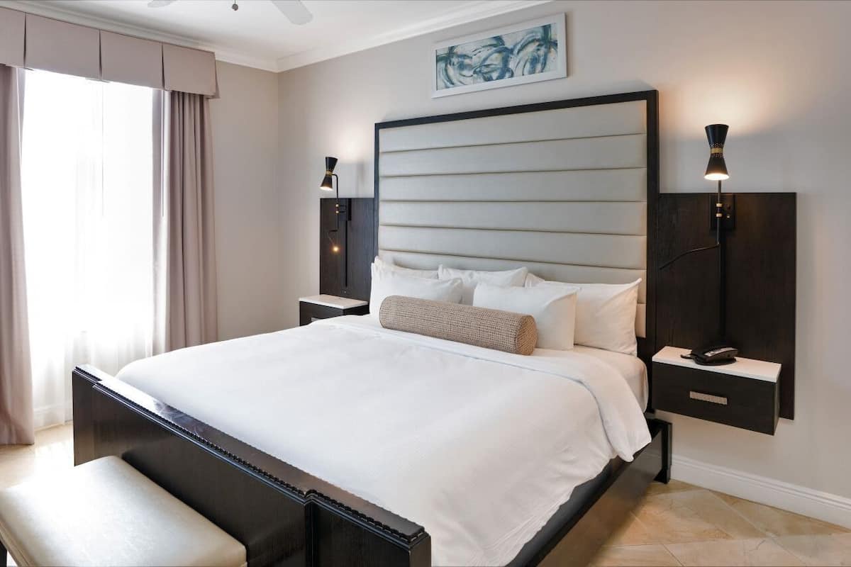 A modern hotel room with a large bed, a bench, a nightstand, and a wall-mounted lamp.