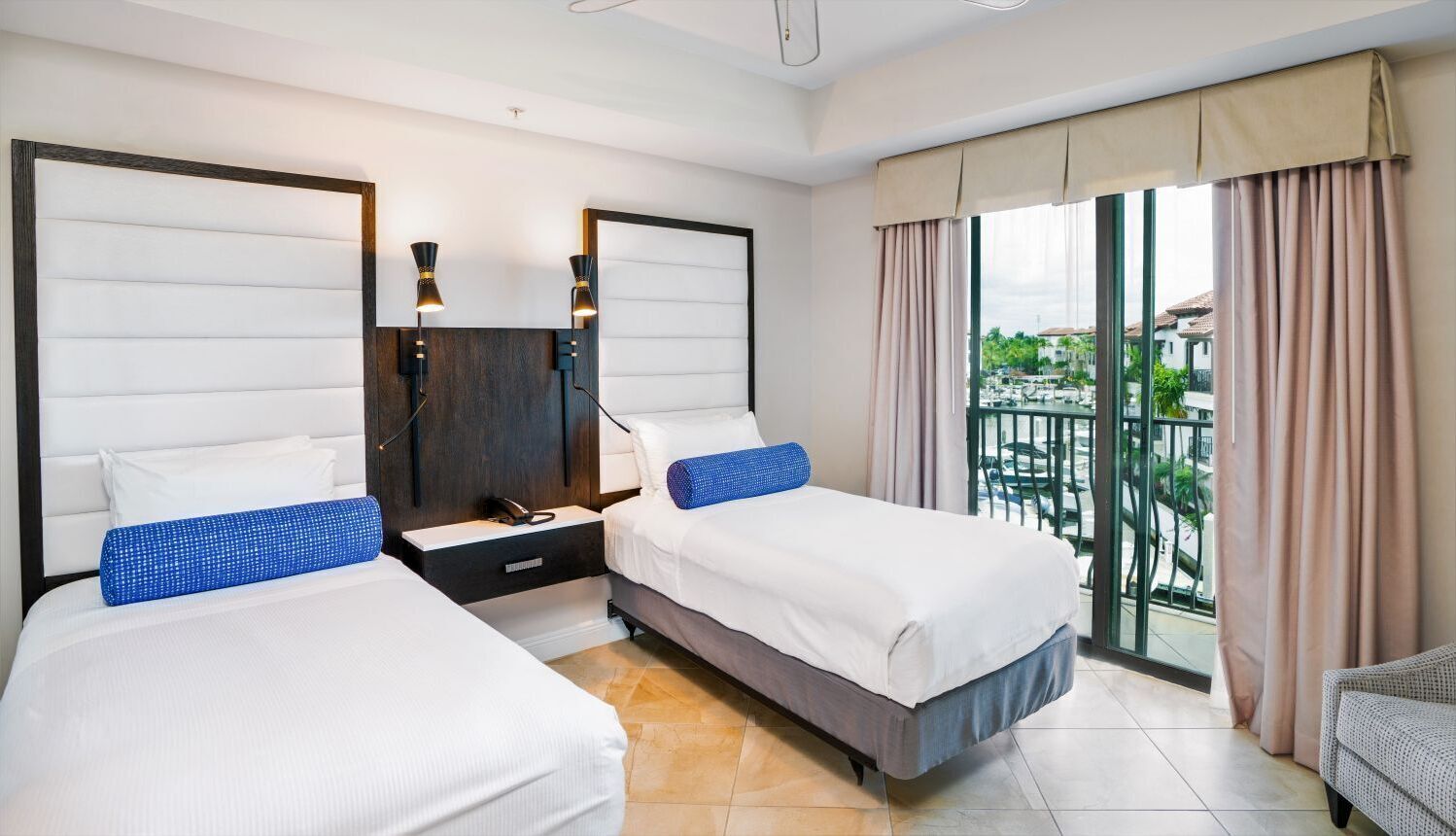 A hotel room with two beds, a balcony with curtains, and a view of the outside.