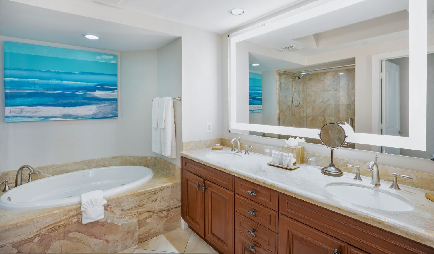 A bathroom with a large bathtub, two sinks, a vanity with drawers, a large mirror, and a painting of the ocean on the wall.