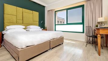 A hotel room with a large bed, a desk with a chair, a window with curtains, and a view of buildings outside.