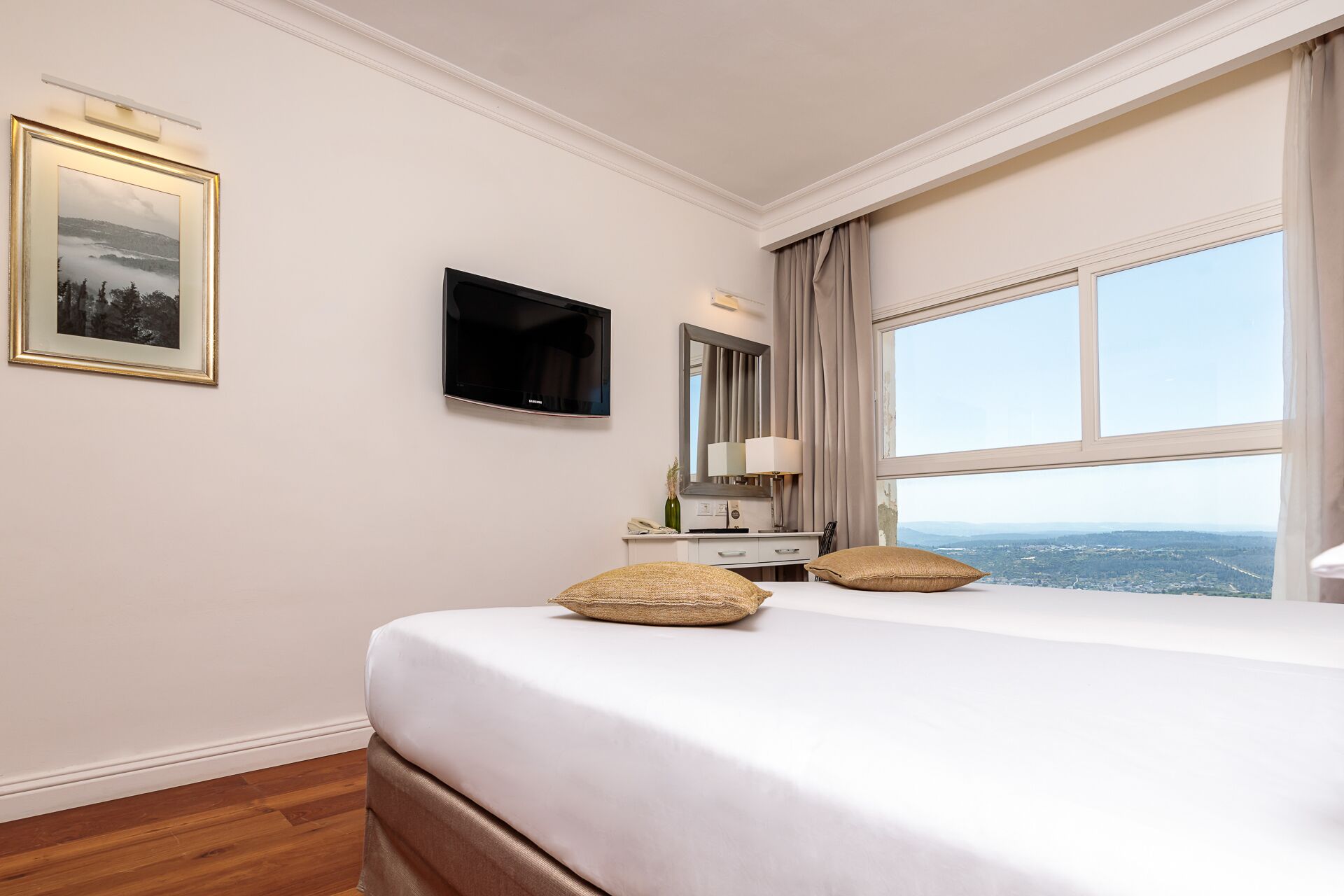 Une chambre d’hôtel dotée d’un grand lit, d’un téléviseur à écran plat, d’une table de chevet avec une lampe, d’un miroir et offrant une vue sur le paysage urbain grâce à de grandes fenêtres.