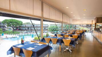 Un amplio comedor interior con mesas y sillas, y grandes ventanales que ofrecen vistas a una piscina y follaje exterior.