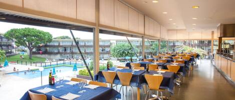 A spacious indoor dining area with tables and chairs, large windows offering views of a pool and greenery outside.