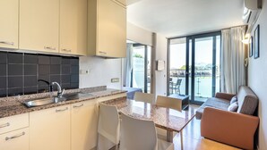 Cocina moderna con encimera de granito, pileta de acero inoxidable y muebles bajos blancos. Incluye un comedor con mesa y sillas, además de un sofá. Amplias ventanas ofrecen vistas al exterior.