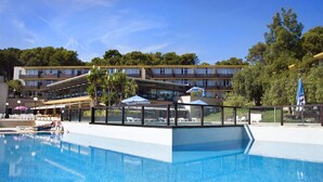 Una piscina con sillones de descanso y sombrillas, rodeada por un edificio con varios balcones y árboles.
