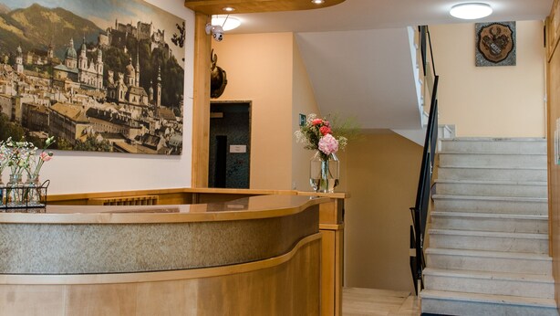 A reception area with a wooden reception desk, a large cityscape painting, a staircase, and a vase with flowers.
