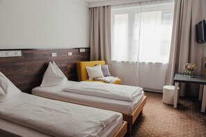 Habitación de hotel con dos camas individuales, cabecera de madera, escritorio pequeño, silla, televisor y ventana con cortinas.