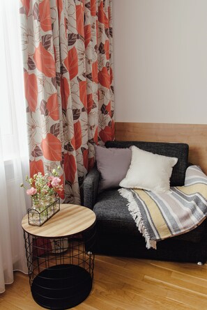 Una habitación con un sofá, una pequeña mesa redonda con un florero y una cortina con estampado de hojas.