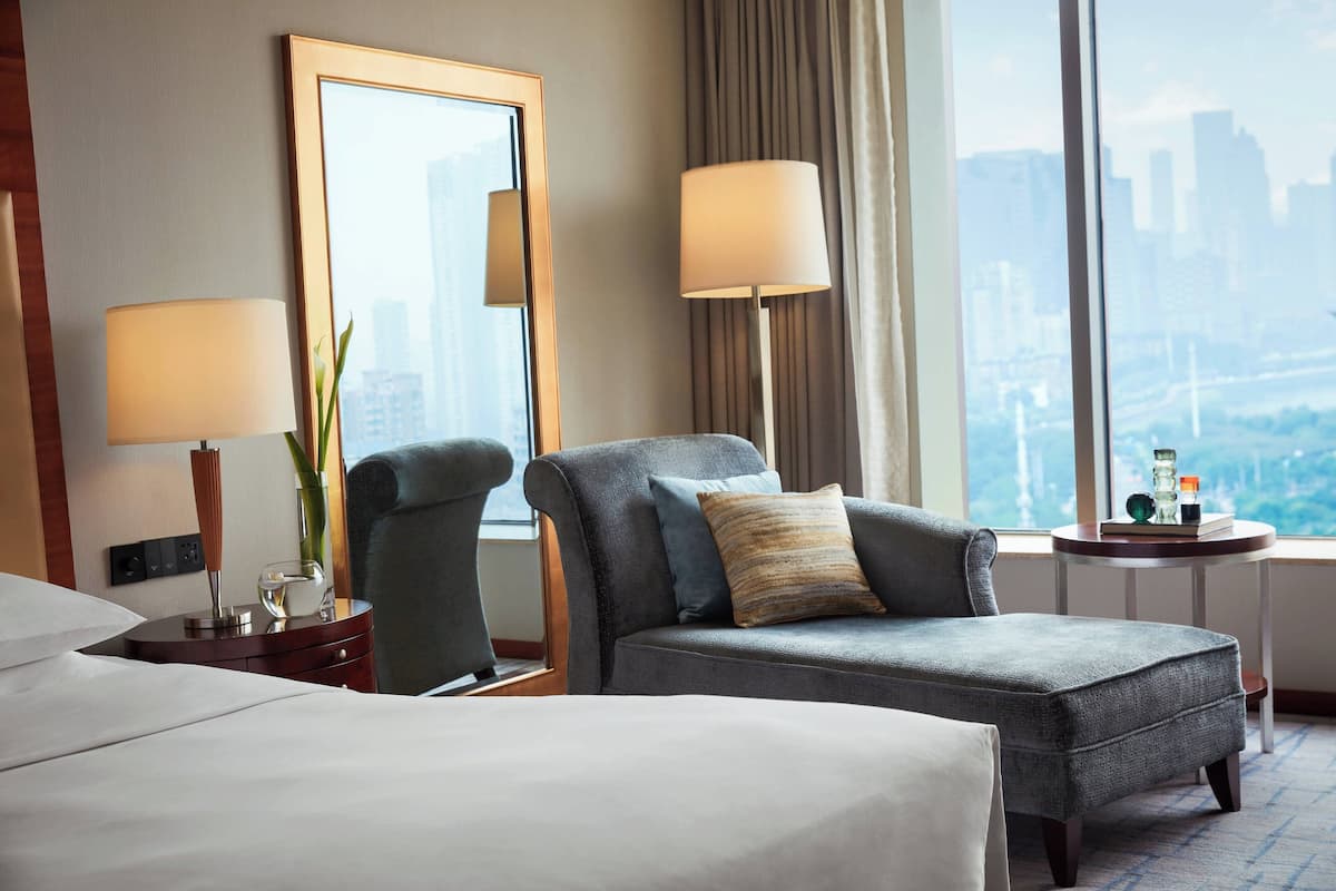 A hotel room with a large bed, two armchairs, a side table with a lamp, a mirror, and a view of the cityscape through large windows.