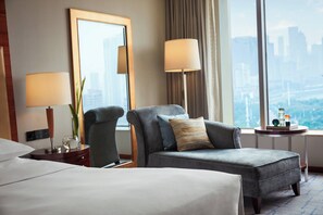 A hotel room with a large bed, two armchairs, a side table with a lamp, a mirror, and a view of the cityscape through large windows.