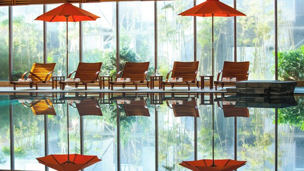 A poolside area with wooden lounge chairs and red umbrellas, reflecting in the water.