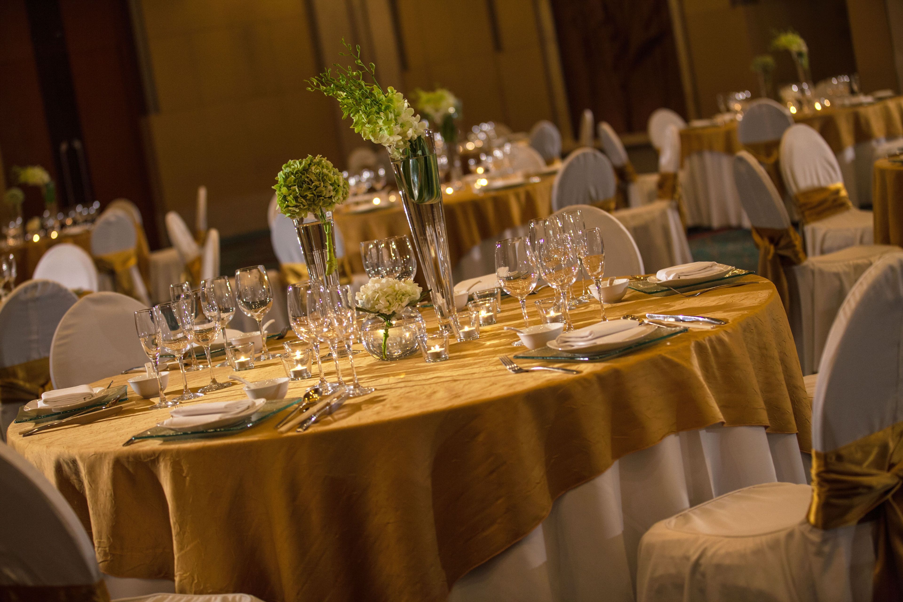 Un salón de banquetes con mesas redondas cubiertas con manteles dorados, sillas blancas y centros de mesa florales.