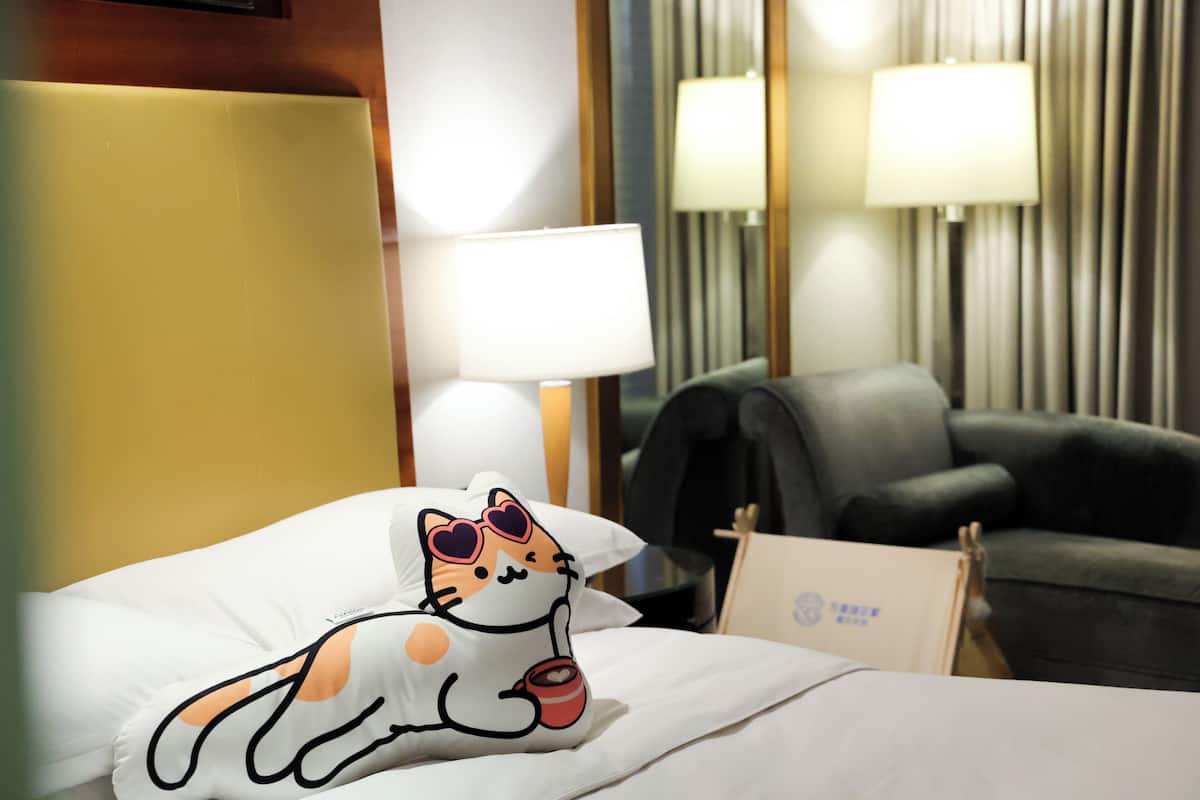 A hotel room with a bed, a pillow with a cat design, a chair, and lamps.