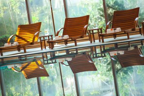 A row of wooden lounge chairs with towels, a glass table, and a view of a forest through large windows.