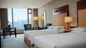 A hotel room with two beds, a desk, and a chair. There is a large window with a view of buildings.