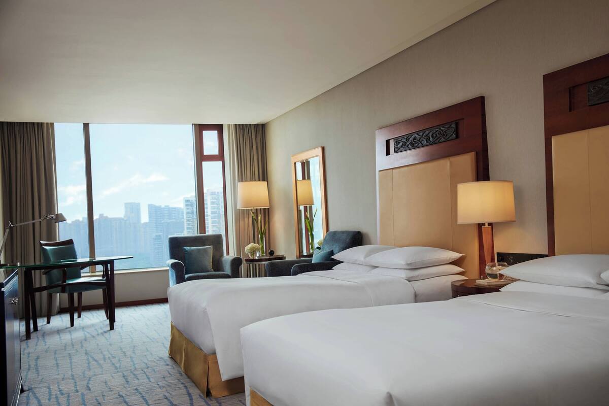 A hotel room with two beds, a desk, and a chair. There is a large window with a view of buildings.