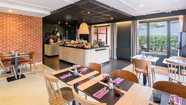A modern dining area with wooden tables and chairs, a central island with kitchen appliances, and large windows offering a view of the outdoors. - Campanile NATURE - Cahors (Cahors)
