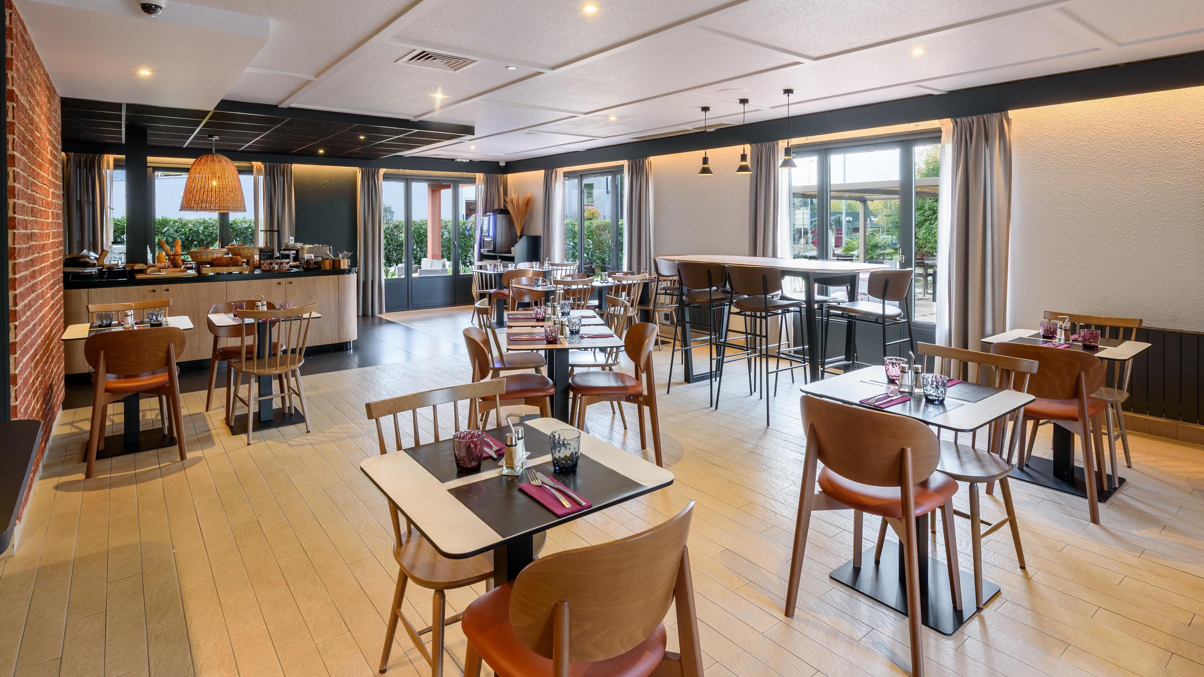 Restaurante moderno com mesas e cadeiras de madeira, área de cozinha e janelas amplas com vista para o exterior.