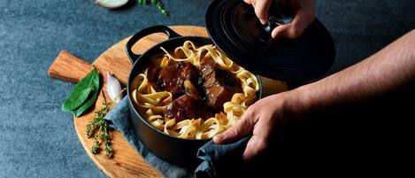 A person lifting a lid off a pot of beef stew with noodles on a wooden board.