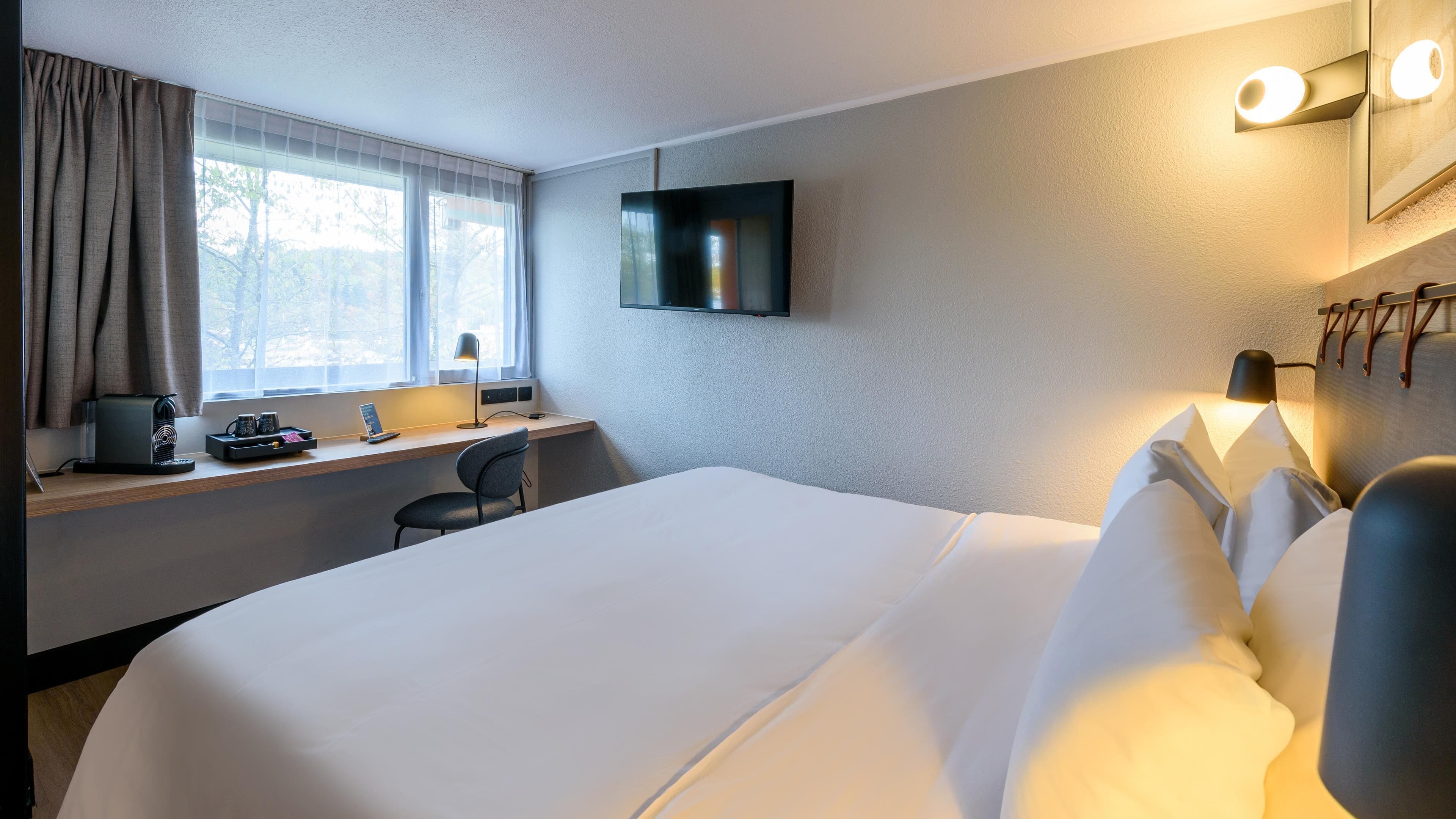 A hotel room with a large bed, a desk with a computer, a chair, a TV mounted on the wall, and a window with curtains.